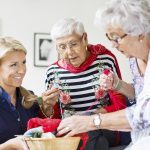 Happy female caretaker looking at senior women choosing wool for knitting at nursing home