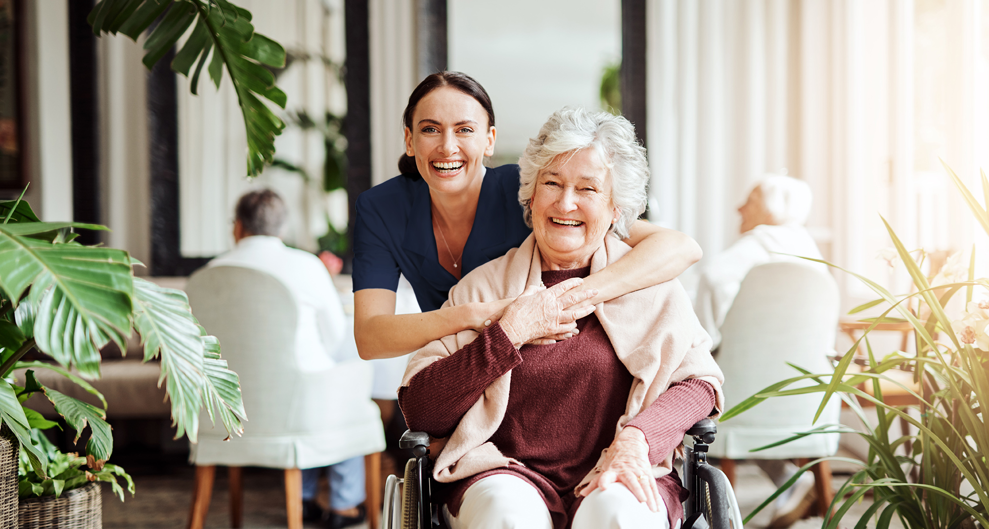 A Support Worker hugging an elderly woman in a wheelchair smiling at the camera